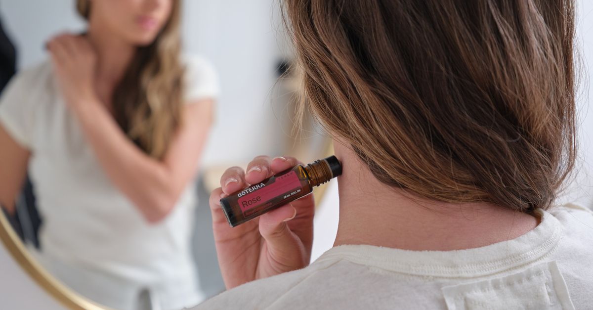 Woman in white shirt applying rose essential oil from roll-on bottle in front of mirror.